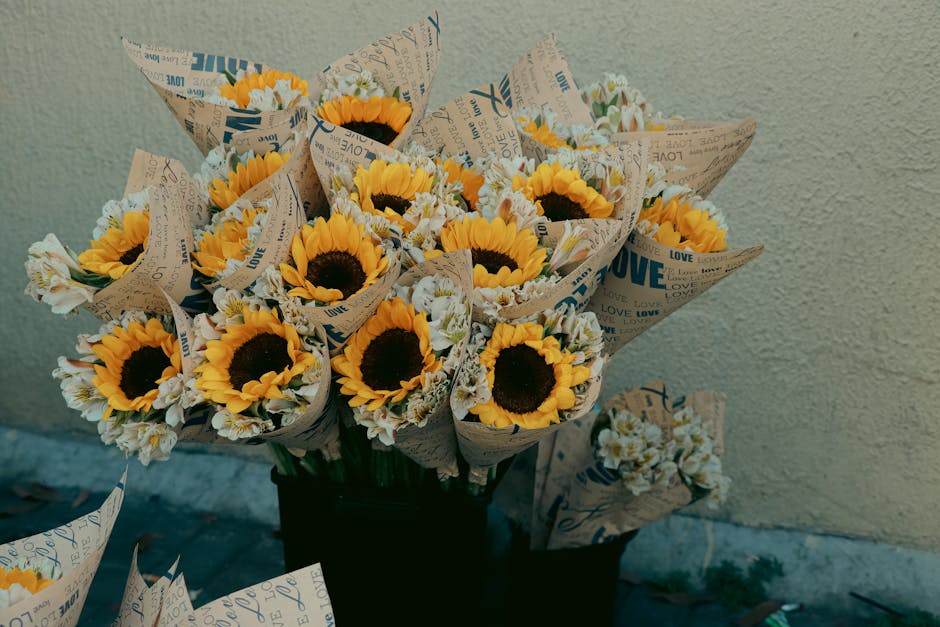 A bouquet of fresh sunflowers with large, vibrant yellow petals and dark brown centers, interspersed with smaller white and pale yellow filler flowers. The flowers are wrapped in rustic brown paper pr