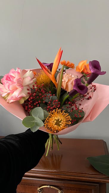 A vibrant floral bouquet featuring a mix of fresh flowers including pink carnations, purple calla lilies, orange and yellow proteas, and Bird of Paradise with an orange bract, wrapped in soft pink tis