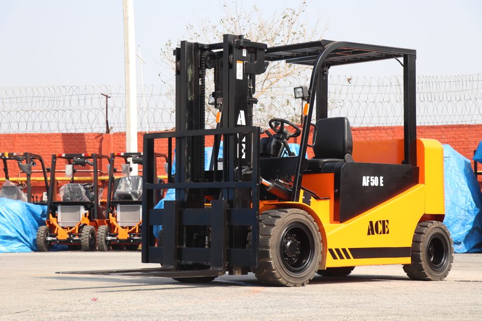 A yellow and black forklift truck with the brand name 'ACE' on its side, parked outdoors on a paved surface. The forklift has a black protective overhead guard, a steering wheel, and a control panel v