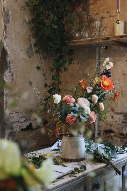A floral arrangement featuring a variety of fresh flowers including blush pink roses, white lisianthus, orange poppies, and dark purple anemones, artistically arranged in a textured, light-colored cer