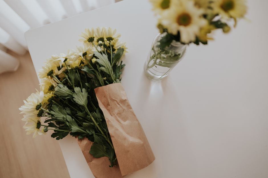 A bouquet of fresh yellow and white daisy-like flowers with green foliage, wrapped in brown kraft paper, resting on a white surface. To the right, there is a glass vase filled with similar yellow and