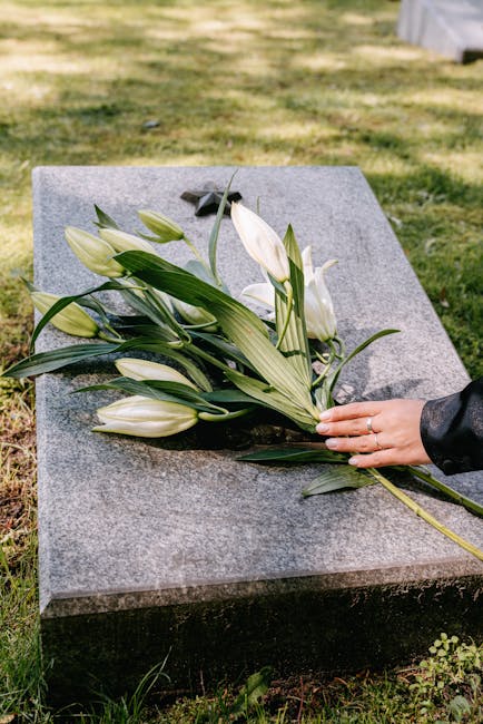 A person's hand, wearing a black leather jacket, gently places a bouquet of white lilies and green foliage onto a polished grey granite tombstone in an outdoor cemetery setting. The bouquet features s
