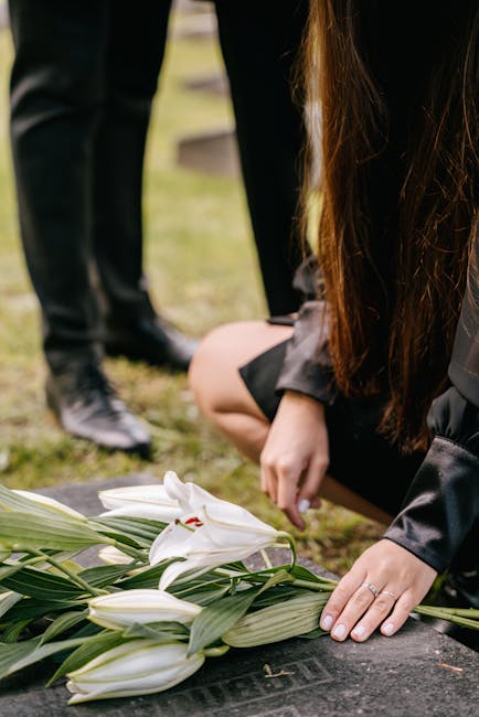 A person wearing a black coat is kneeling on the ground, gently placing a floral arrangement of white lilies with green leaves on a gravestone. The lilies are fresh, with open petals revealing red sta