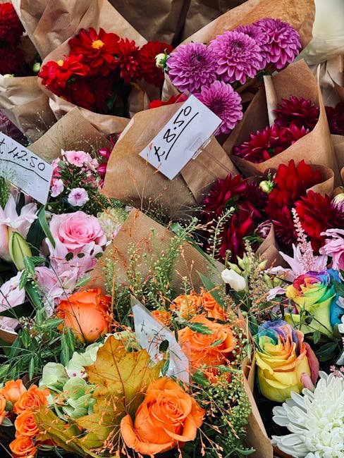 A colorful assortment of fresh flowers arranged in brown paper wrapping, including pink and purple chrysanthemums, vibrant red dahlias, peach roses, rainbow-hued roses, and white blooms, all displayed